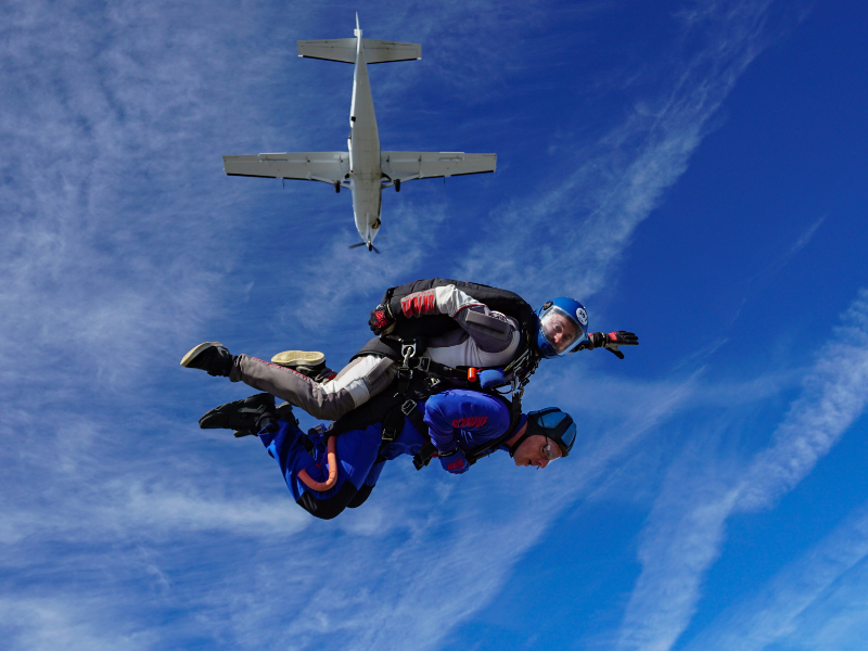An image of someone doing a tandem skydive against bright blue skies and the plane they jumped from above.