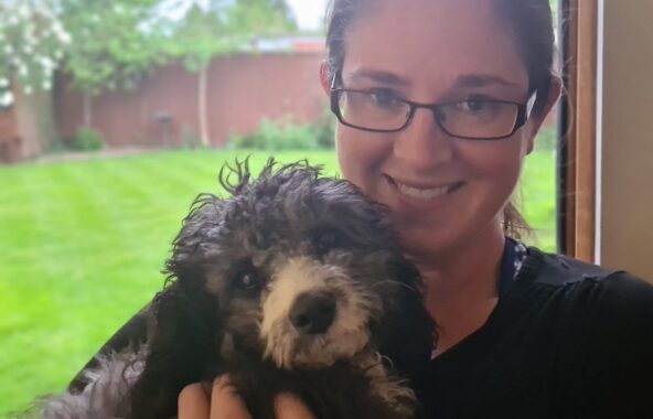 Lady in twenties on right hand side of image with light brown hair tied back wearing rectangular framed black glasses and a black t-shirt. Holding a black and white poodle cross dog and a garden can be seen in the background