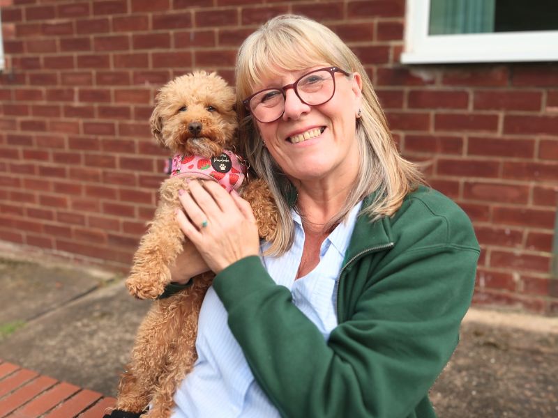 Middle-aged women with light blonde/grey hair and glasses wearing a green open jumped and blue shirt holding a small ginger curly dog wearing a pink harness.