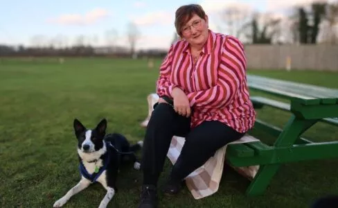 Middle aged lady with cropped hair wearing a pink striped shirt and black trousers sat on a green bench on a field with a black and white Collie dog laid at her feet