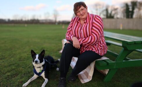 Middle aged lady with cropped hair wearing a pink striped shirt and black trousers sat on a green bench on a field with a black and white Collie dog laid at her feet