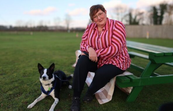Middle aged lady with cropped hair wearing a pink striped shirt and black trousers sat on a green bench on a field with a black and white Collie dog laid at her feet