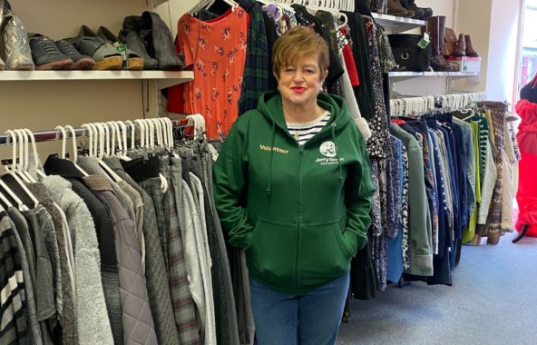 A brunette woman standing infront of clothings rails in a charity shop. She is wearing a green Jerry Green Dog Rescue Volunteer jumper.