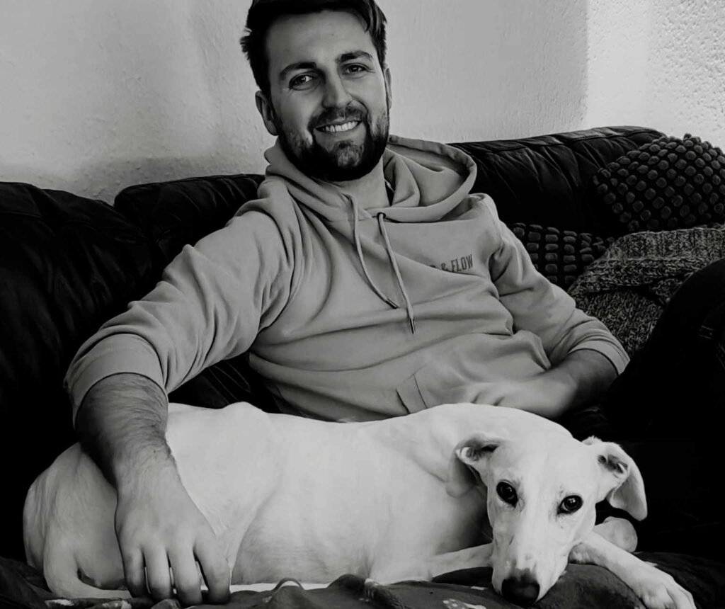 Black and white photo of a man in his late twenties to earlier thirties with dark hair and a short dark beard wearing what appears to be a grey hoodie sat on a sofa with a white Lurcher dog curled up next to him looking directly at the camera. The man is smiling and his arm is over the dog.