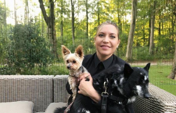Blonde lady with hair tied back sat on grey rattan furniture with woods behind her. In her arms are a senior Yorkshire Terrier dog and a black and white cross breed dog