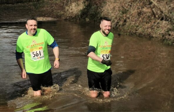 Two middle-aged men wearing neon green Go Extreme with Jerry Green wading through muddy water in a fundraising challenge