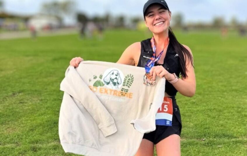 Young woman wearing a black vest, shorts and cap with long dark hair in a ponytail holding a Go Extreme with Jerry Green cream hoodie in front of her smiling at the camera after completing a fundraising challenge