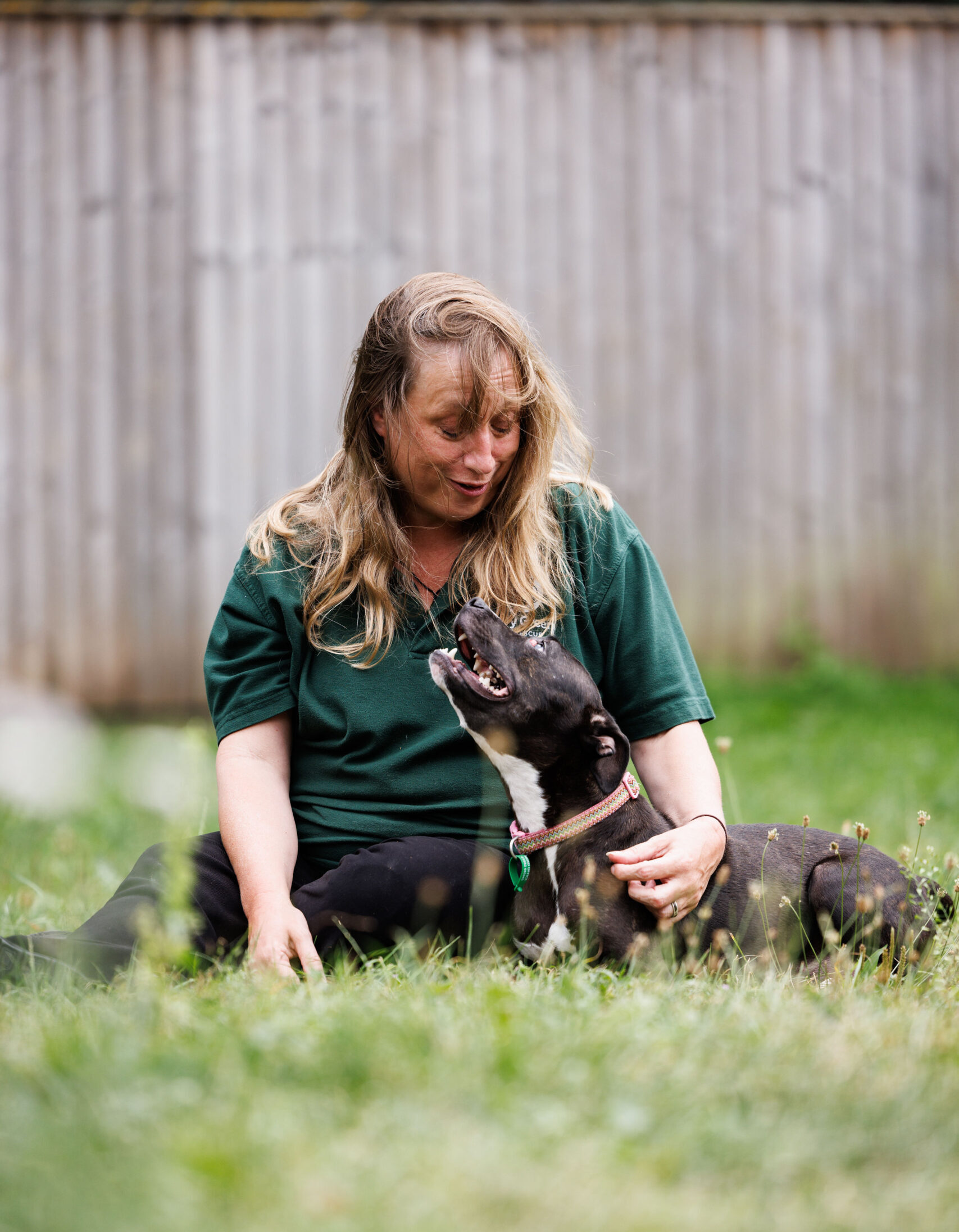 A blonde woman wearing a green tshirt, sitting in a garden whilst looking down at a Staffordshire Bull Terrier. The dog is laying down, and wearing a pink collar whilst looking up at the woman.