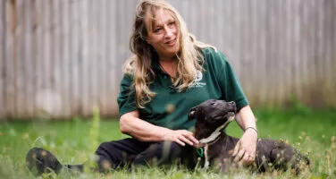 Blonde Jerry Green Dog Rescue staff member sat in grass with black staffordshire bull terrier