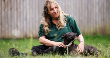 Blonde Jerry Green Dog Rescue staff member sat in grass with black staffordshire bull terrier