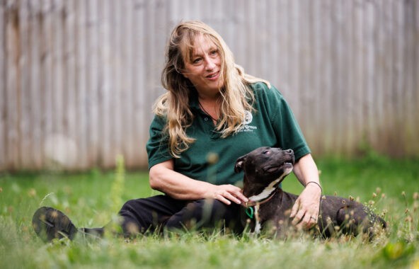 Blonde Jerry Green Dog Rescue staff member sat in grass with black staffordshire bull terrier
