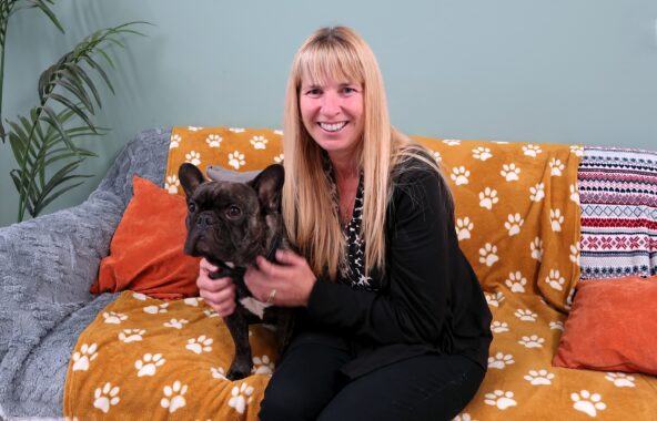 Blonde middle-aged lady sat with a brindle French bulldog on a sofa covered in multiple different coloured blankets on a light teal background with a plant in the left corner
