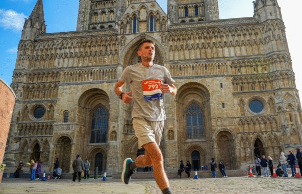 Middle-aged man wearing grey t-shirt and shorts with dark grey socks and trainers wearing a marathon ballot number on his chest running in front of Lincoln Cathedral