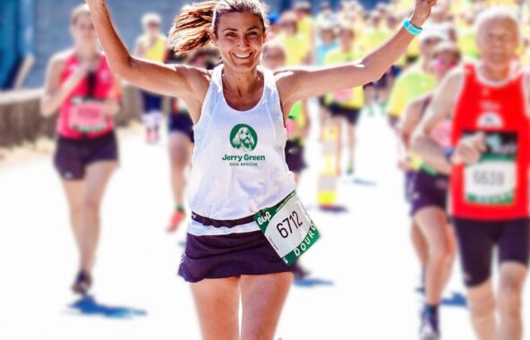 Woman running with brown hair in pony tail wearing white Jerry Green Dog Rescue vest and black shorts with marathon ballot number holding arms up with hands in peace signs