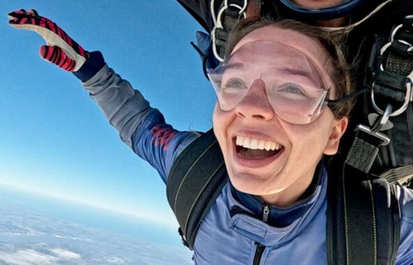 Young female with blonde hair tied back doing a skydive wearing a blue jumpsuit and red gloves, goggles and black harness with skydiving instructor cut out of photo above