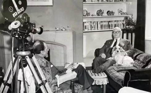 Black and white photo of a man in the 1960s sat on a sofa with a Cocker Spaniel being interviewed by a man sat opposite with a film reel camera behind him
