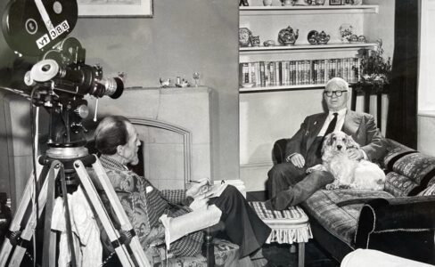 Black and white photo of a man in the 1960s sat on a sofa with a Cocker Spaniel being interviewed by a man sat opposite with a film reel camera behind him