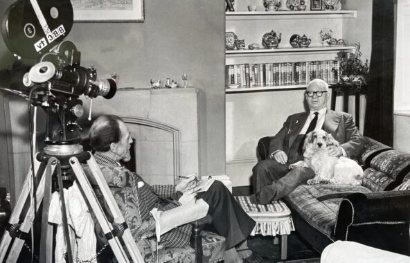Black and white photo of a man in the 1960s sat on a sofa with a Cocker Spaniel being interviewed by a man sat opposite with a film reel camera behind him