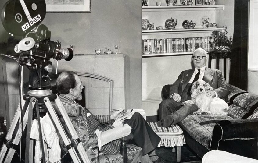 Black and white photo of a man in the 1960s sat on a sofa with a Cocker Spaniel being interviewed by a man sat opposite with a film reel camera behind him