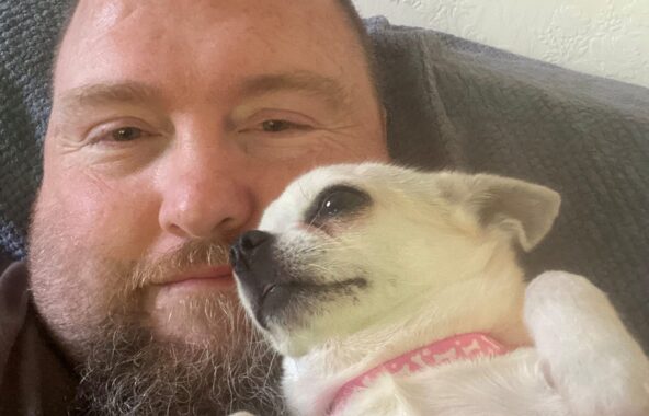Happy looking cream chihauha lying tummy upwards on the chest of a man with a beard.
