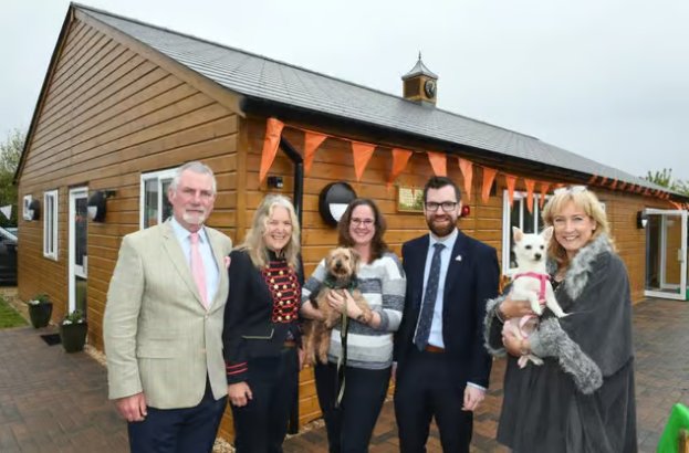 Five individuals stood in front of a new building with orange bunting with two women holding two small white dogs