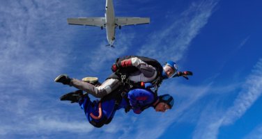 An image of someone doing a tandem skydive against bright blue skies and the plane they jumped from above.