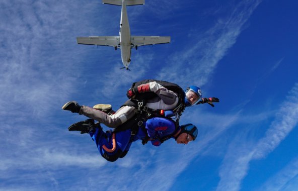 An image of someone doing a tandem skydive against bright blue skies and the plane they jumped from above.