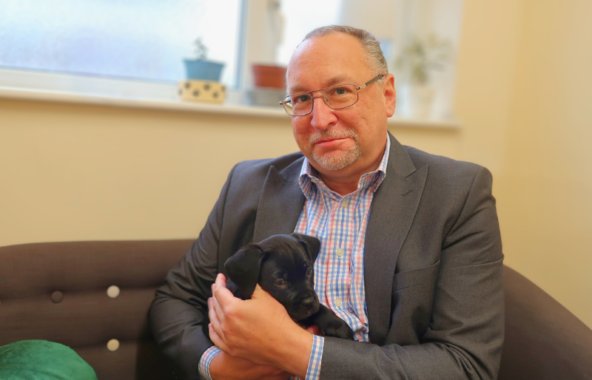 Man with dark grey hair and goatee wearing silver framed glasses and a grey suit with a finely checked shirt holding a black Cane Corso puppy on is lap on a brown sofa with a green cushion on the left