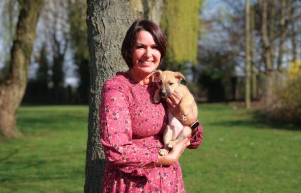 Woman in a pink dress standing in front of a tree holding a small cream coloured dog.
