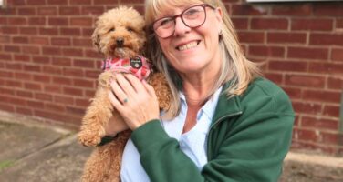 Middle-aged women with light blonde/grey hair and glasses wearing a green open jumped and blue shirt holding a small ginger curly dog wearing a pink harness.
