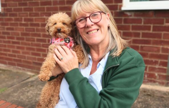 Middle-aged women with light blonde/grey hair and glasses wearing a green open jumped and blue shirt holding a small ginger curly dog wearing a pink harness.