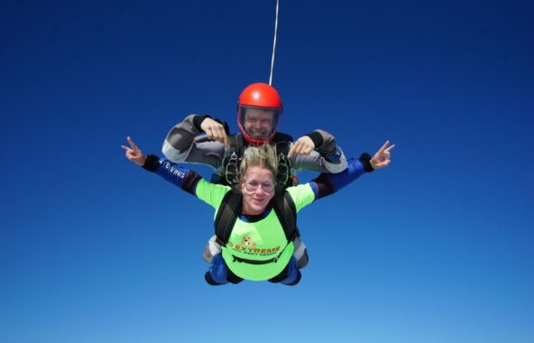 Tandem skydivers, man with red helmet and grey suit, young woman with yellow and blue top and hair swept back by wind.